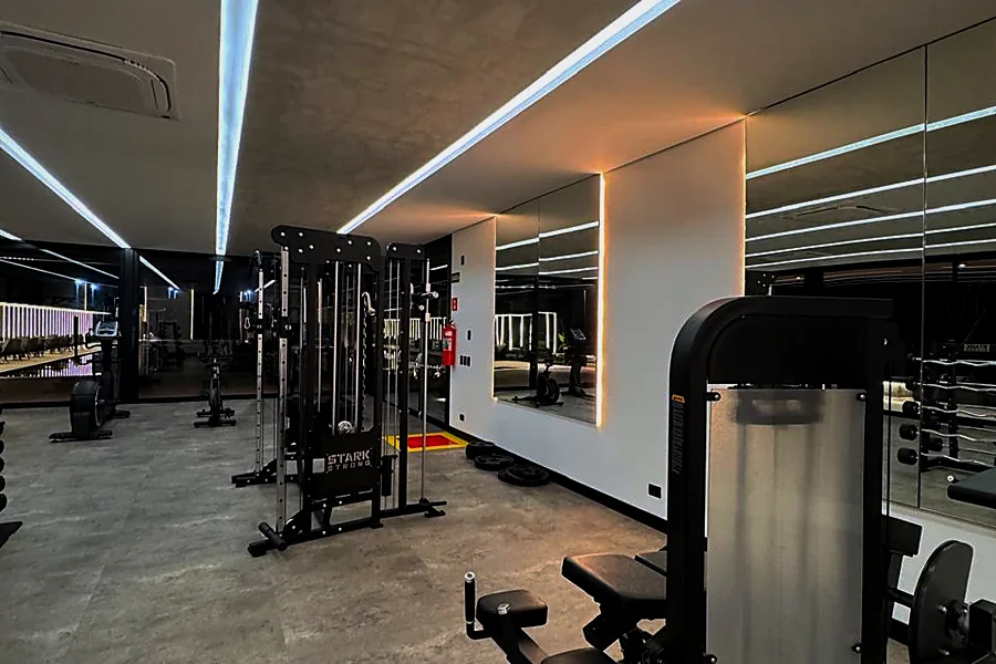 Academia do clube do Infinitus, com estação de musculação, espelhos e iluminação linear no teto